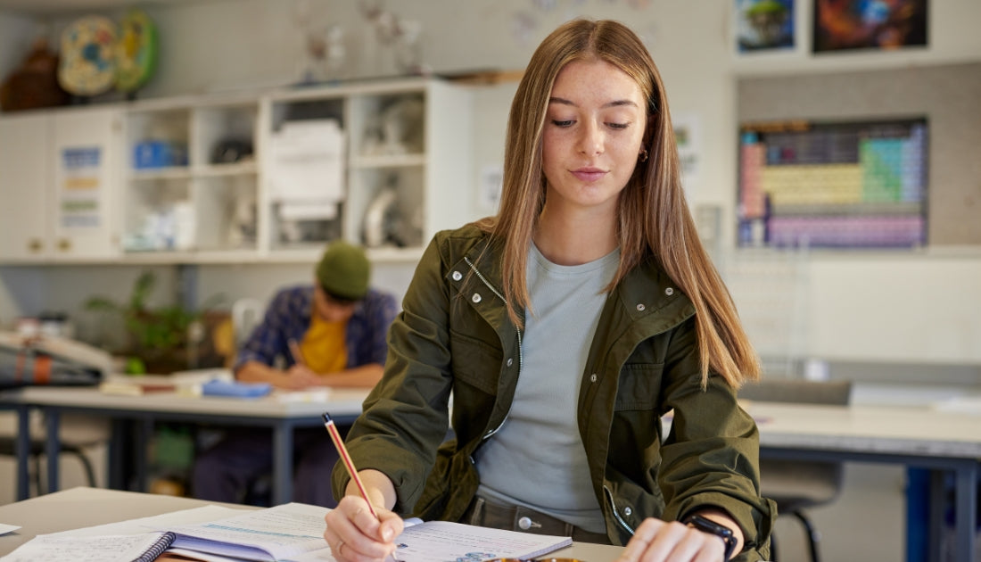 A student sits at a desk in a classroom, writing in a notebook while another student works in the background. Science posters and classroom shelves are visible behind them.